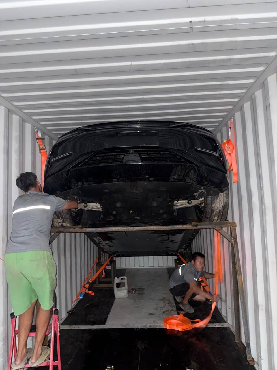 Two workers strapping a vehicle onto a frame inside a container